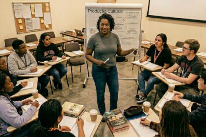 Professora conduz aula de produção cultural com jovens estudantes sentados em círculo em sala de formação, com quadro ao fundo e anotações sobre análise crítica e projetos culturais.