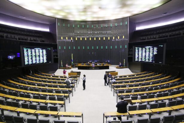 Plenário da Câmara dos Deputados com bancadas curvas, muitas cadeiras vazias, poucas pessoas e dois painéis eletrônicos laterais.